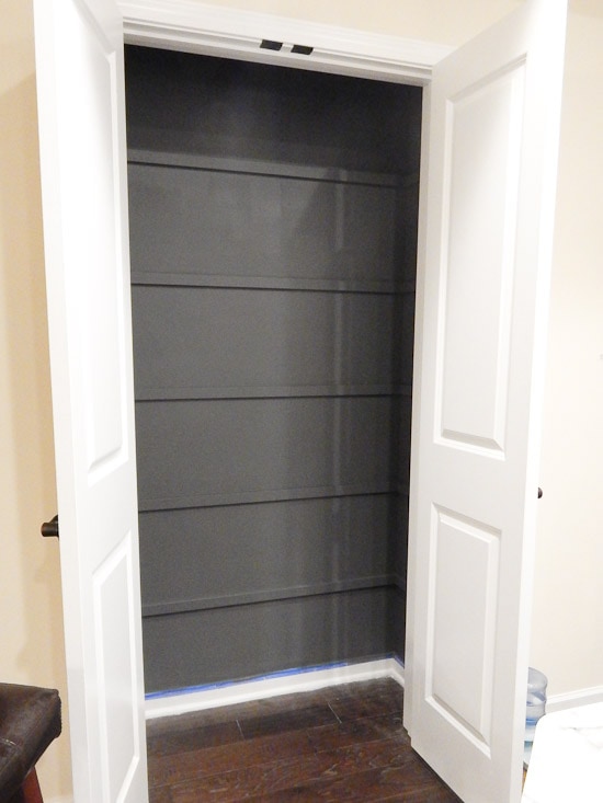 An empty closet with double white doors, slightly ajar, revealing a dark grey back wall and interior shelving ideal for those looking to build pantry shelves. The closet is situated on a wooden floor.