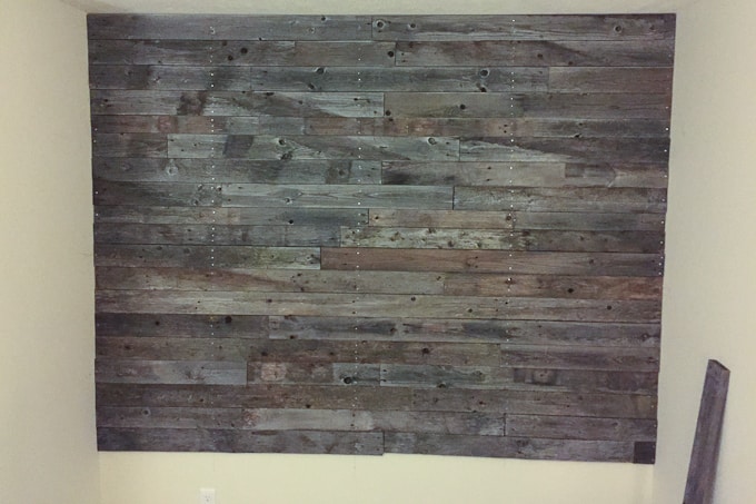 A large wall covered with reclaimed wooden planks of varying shades of brown and gray, arranged horizontally. Creating a custom pallet wall like this contrasts with the surrounding plain beige walls, adding a rustic and textured appearance to the room.
