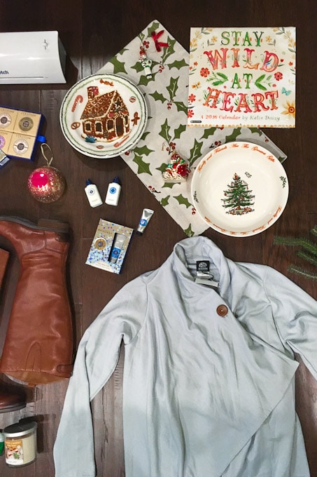 A wooden table displays a collection of holiday items. These include a festive Christmas calendar, holiday-themed plates, a gingerbread house, boots, a grey jacket, and small gift boxes. A small decorated Christmas tree and greeting cards add to the seasonal feel.