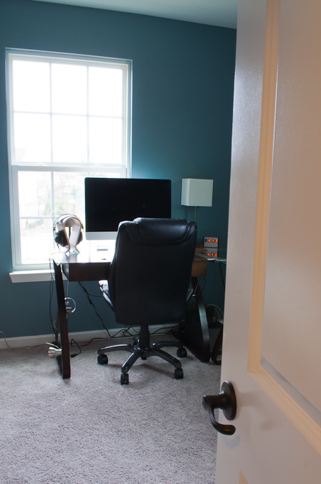 A home office with a large window providing natural light. A desk with a computer monitor, keyboard, and a lamp sits against the wall. A black office chair is positioned in front of the desk, and various cords are visible under the desk. The office door is partially open.