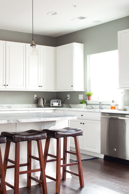 A modern kitchen features white cabinets, a marble countertop, and a stainless steel dishwasher. A pendant light hangs above the island, which is accompanied by two wooden stools with dark cushions. A toaster, kettle, and plant add touches of functionality and decor to the inviting space.