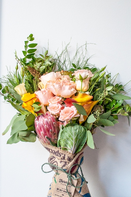 A vibrant bouquet from Farmgirl Flowers featuring a mix of pink roses, yellow calla lilies, greenery, and a single pink protea. The flowers are wrapped in rustic burlap with a visible tag attached, all against a plain light-colored background.