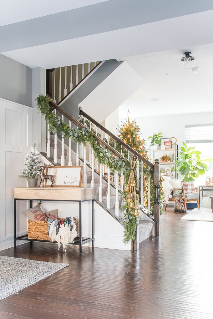 A cozy, festive entryway decorated for the holidays features a wooden staircase adorned with greenery. Below the stairs is a small table with holiday decorations, including a mini Christmas tree, a framed "Merry & Bright" sign, and a woven basket with blankets—perfect for your Modern Classic Christmas Home Tour.