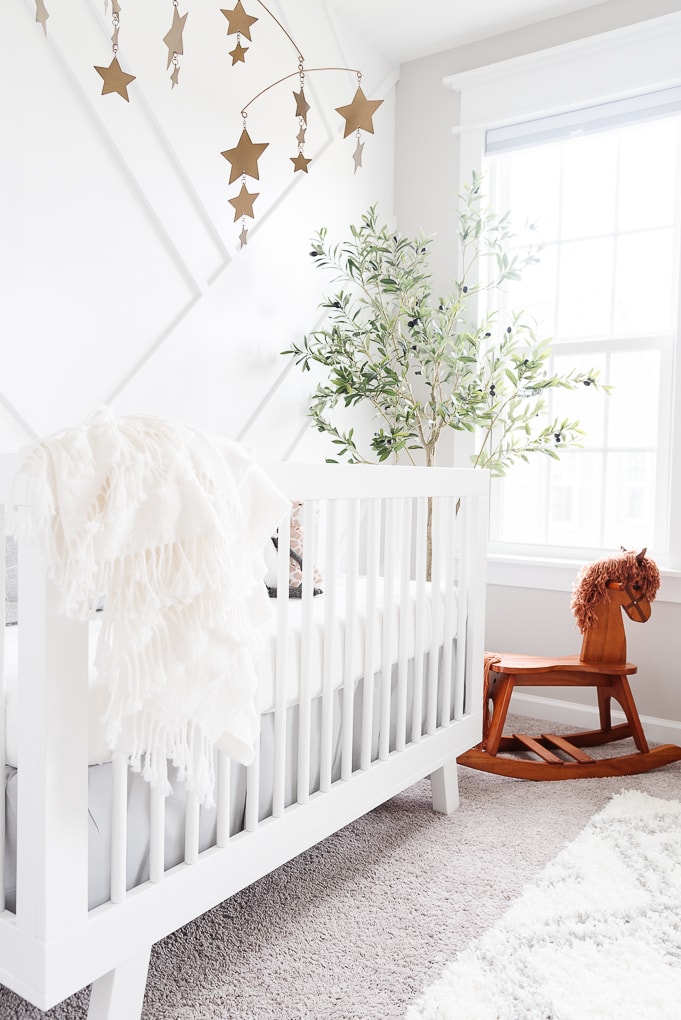 wooden rocking horse next to a white crib in a nursery with a faux olive tree in the background