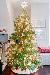 In a cozy Christmas living room, a decorated tree stands in a corner, adorned with various ornaments and bright white lights. A glowing star tops the tree. To the right, a festive red pillow with white "falalalala" text rests on a white woven basket, and nearby, bells hang from a banister.