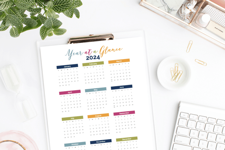 Top view of a Year at a Glance Calendar 2024 on a clipboard. The desk includes a plant, keyboard, cup of coffee, paper clips, and various desk accessories. The calendar features each month's dates in a grid, with each month color-coded for easy reference.