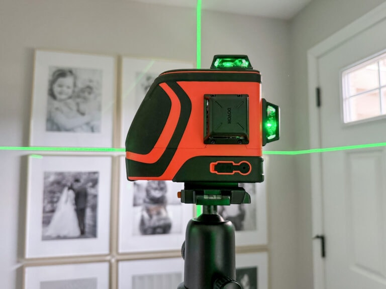 A best budget laser level device on a tripod projects green laser lines horizontally and vertically across a room. The background features framed photographs on a wall and a door with a window.