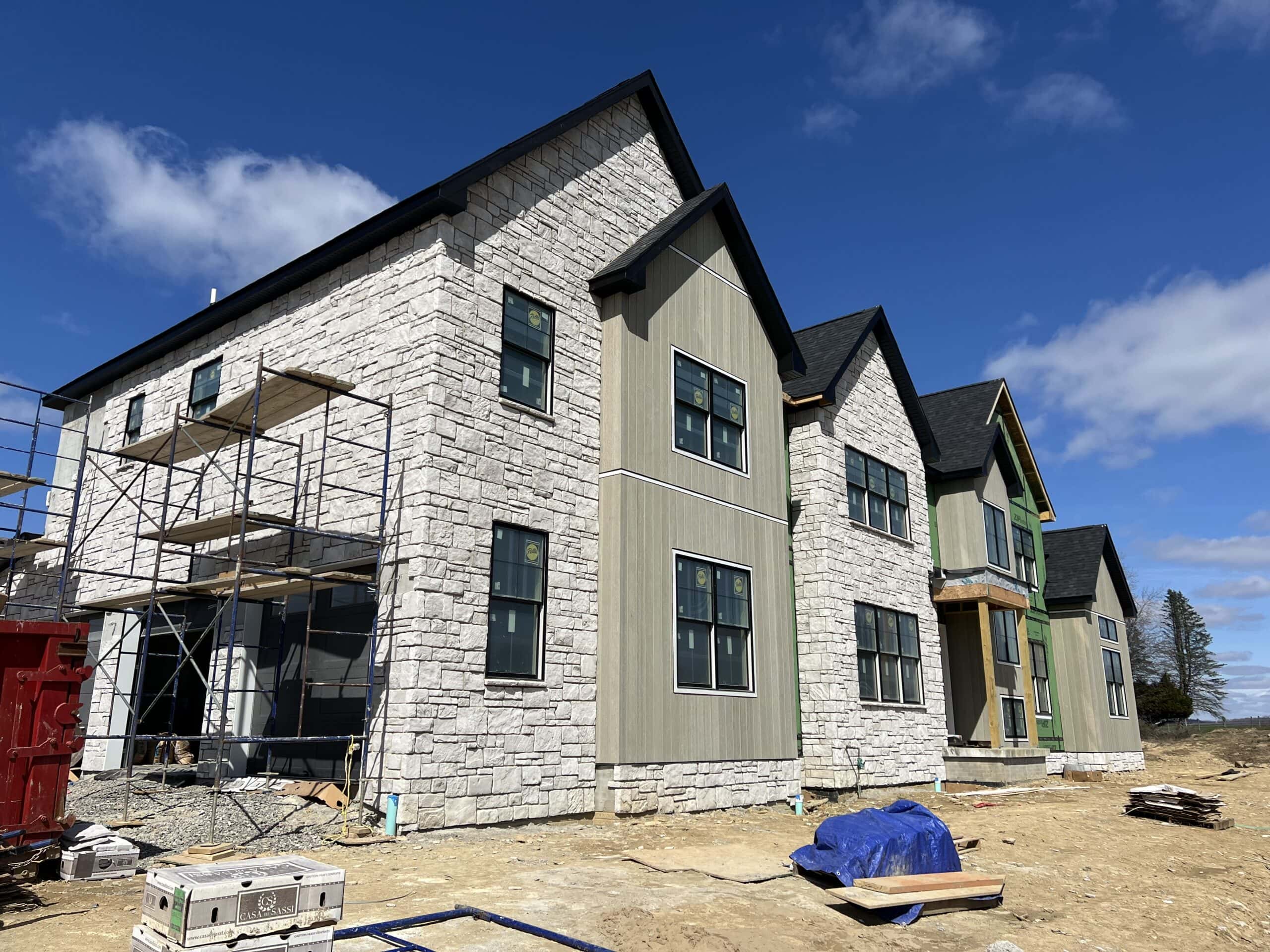 Partially constructed two-story house with a stone facade and green siding, showcasing the custom home building journey. Scaffolding is set up on one side, and construction materials are scattered around the site. The sky is clear and blue, and there are a few trees in the background.