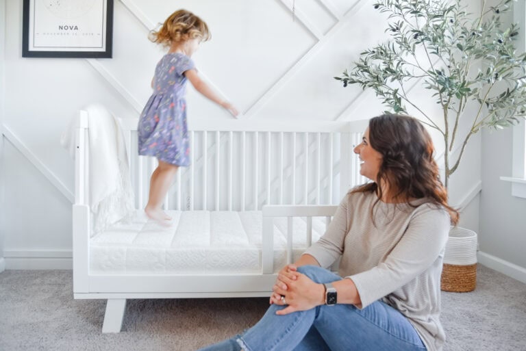 A woman with long brown hair sits on the floor, smiling at a young girl in a floral dress who is jumping on a white toddler bed. The room, part of their clean and organized home with kids, features light-colored walls with decorative molding, a celestial-themed framed print, and a large potted plant.