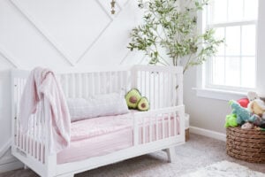 A cozy, clean, and organized nursery features a white crib with light pink bedding and a gray pillow. A pink blanket drapes over the crib's side. Decorative pillows shaped like avocados sit inside. A green plant stands by the window, and a basket filled with stuffed animals is on the floor.