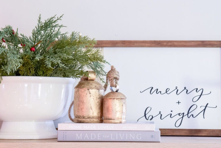 A festive display seamlessly capturing the transition from fall to winter features a white pot of greenery and red berries, rustic metal bells, and a wooden frame with "merry + bright." Two stacked books titled "Home Made Lovely" and "Made for Living" complete this cozy home decor.