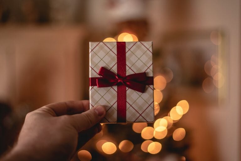 A hand holding a small, carefully organized gift wrapped in patterned paper with a red bow. Warm, blurry lights create a cozy, simplifying festive background perfect for the holidays.