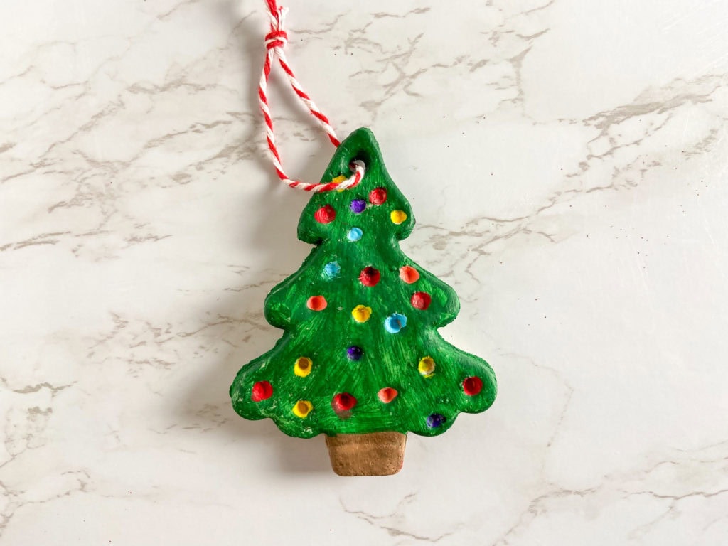 A handmade Christmas tree ornament, painted green with colorful dots resembling baubles, hangs from a red and white striped string. Perfect for Christmas crafts sessions, it stands out against the white marble surface, creating holiday memories that last a lifetime.