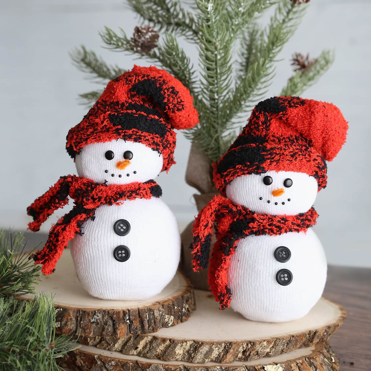 Two handmade snowmen with red and black hats and scarves stand on stacked wooden logs, evoking warm holiday memories. They have button eyes, carrot noses, and black buttons on their bodies. In the background, green pine branches add a festive touch to this kid-friendly scene.