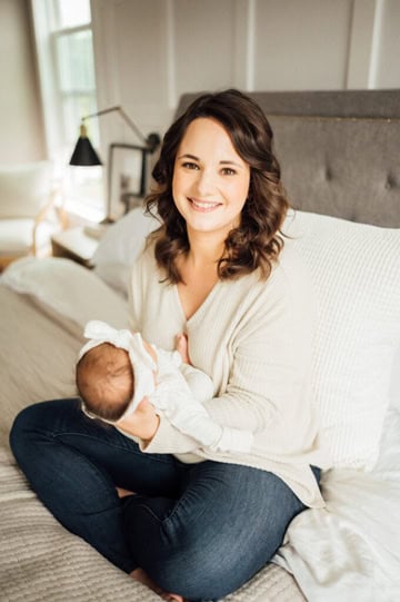 A woman with brown hair sits cross-legged on a bed, smiling at the camera while gently cradling a baby dressed in white. The room is softly lit, with neutral decor and a cozy atmosphere.